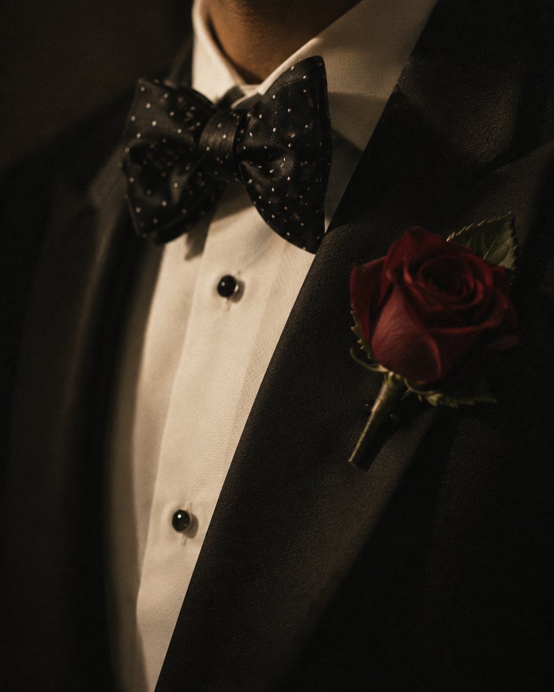 A black polka-dot bow tie on a tuxedo, with a deep red rose boutonnière